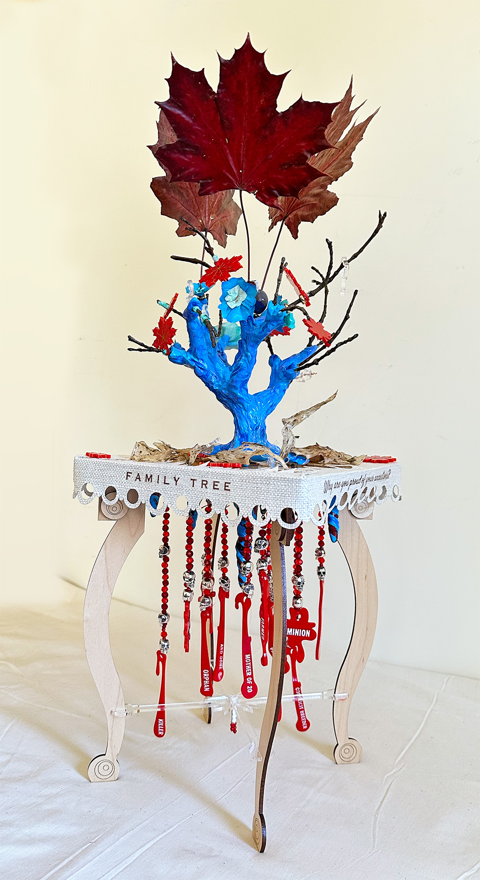 A 3D multi-media piece which depicts a maple wood table on the top grows a blue tree trunk with some bare branches and red leaves. Underneath the table top hang the roots and drops of blood suspended by strings of red beads from the roots. There are words on the droplets like; rapist, enslaver, mother of 12, patriarch, killer, soldier, farmer.
